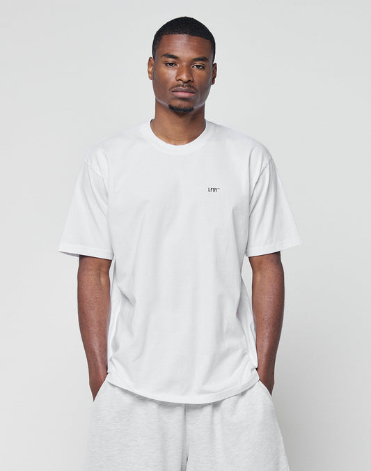A man wears the LIVE FAST DIE YOUNG Basic Chest Tee in plain white and light gray shorts, standing against a simple light gray background with his hands in his pockets, looking straight at the camera.