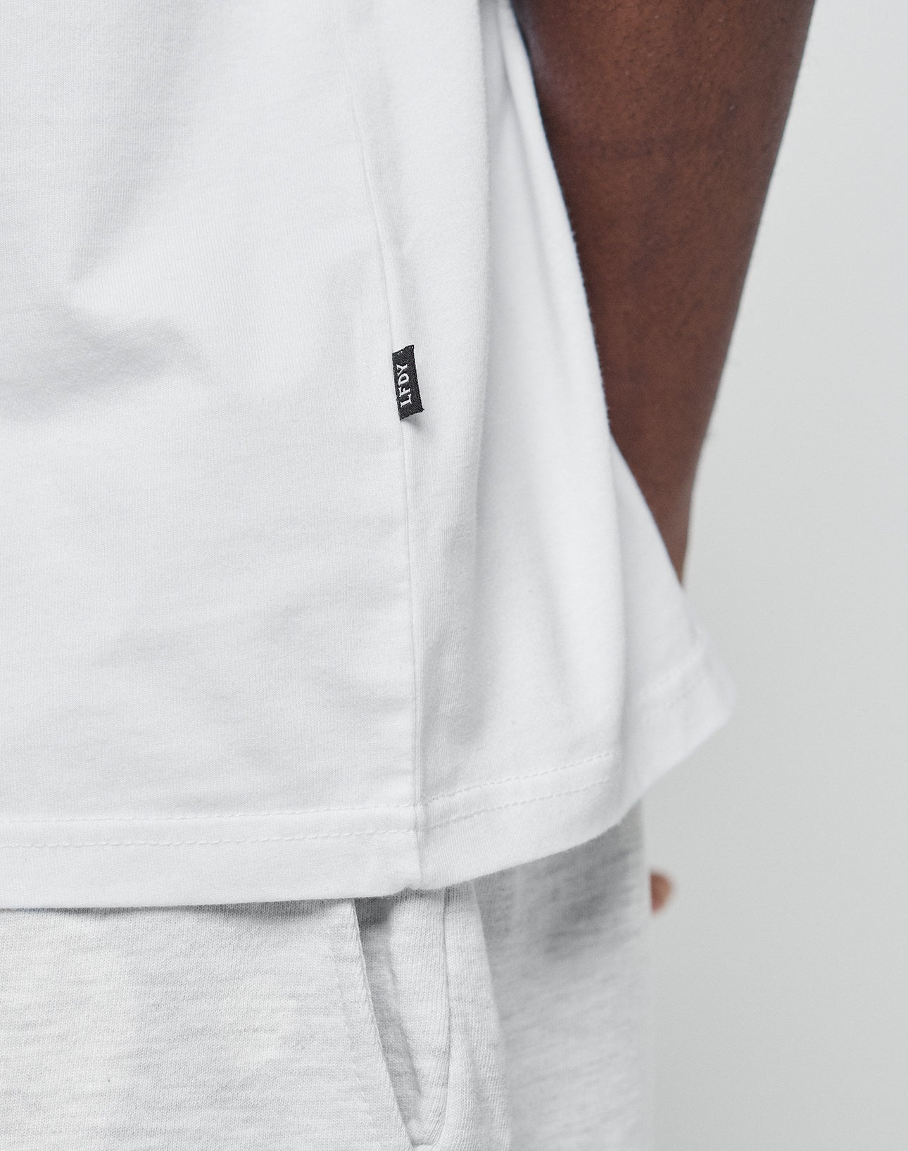 Close-up of a person wearing the LIVE FAST DIE YOUNG Basic Chest Tee in white, made from 100% Baumwolle, paired with light gray pants. The tee features a small black side tag; the persons arm is partly visible against a plain background.