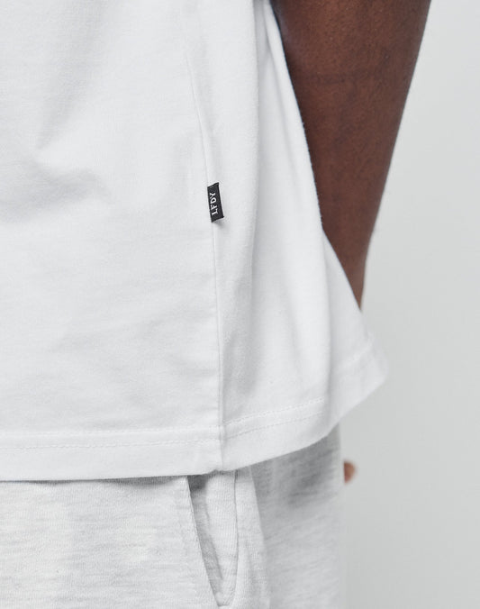 Close-up of a person wearing the LIVE FAST DIE YOUNG Basic Chest Tee in white, made from 100% Baumwolle, paired with light gray pants. The tee features a small black side tag; the persons arm is partly visible against a plain background.