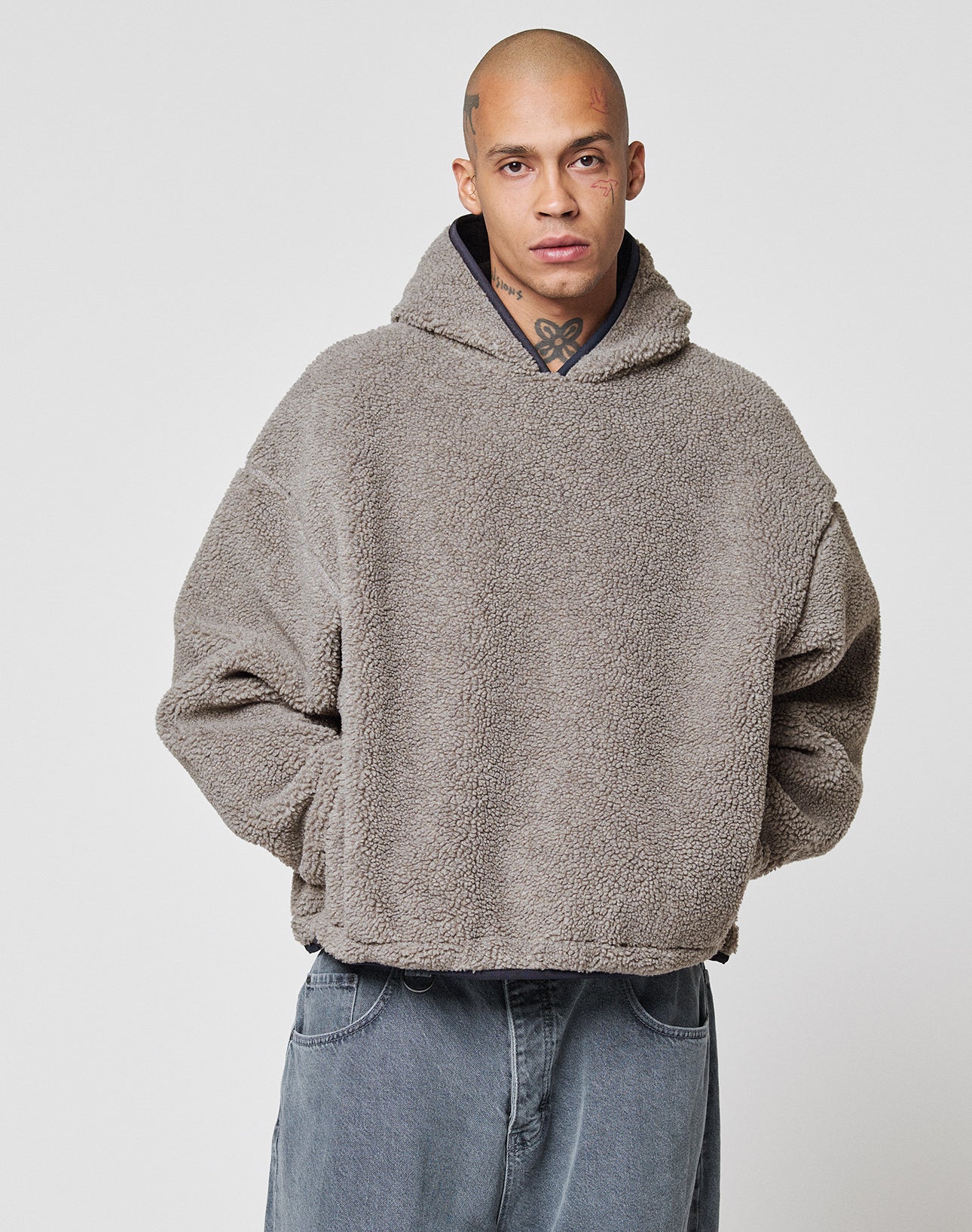 A person with a shaved head and neck tattoos wears the LFDY Teddy Hooded by LIVE FAST DIE YOUNG, an oversized gray fuzzy hoodie with Eingrifftaschen, paired with baggy denim jeans. They stand against a plain white background, hands in pockets.