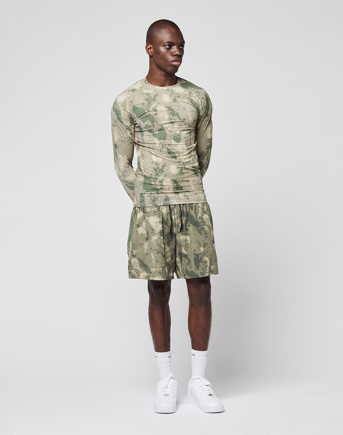A man stands against a plain background wearing the LIVE FAST DIE YOUNG LFDY Performance Slim Longsleeve in green camouflage, with matching shorts, white crew socks, and white sneakers. His arms are behind his back as he looks to the side.