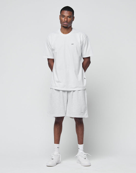 A man stands against a plain white background, facing forward with his hands behind his back. He wears a loose white T-shirt, LIVE FAST DIE YOUNG Basic Sweat Shorts in light gray with an elastic waistband, white socks, and sneakers.