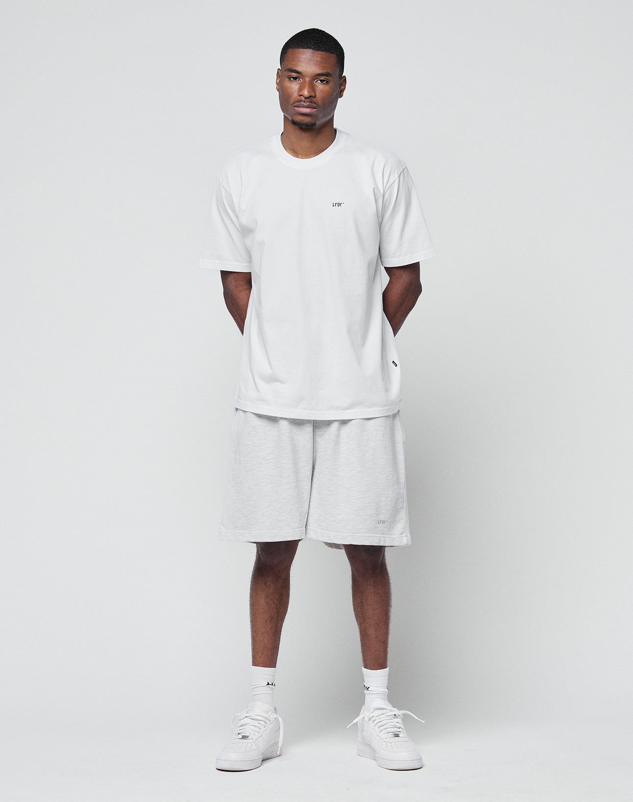 A man stands against a plain white background, facing forward with his hands behind his back. He wears a loose white T-shirt, LIVE FAST DIE YOUNG Basic Sweat Shorts in light gray with an elastic waistband, white socks, and sneakers.
