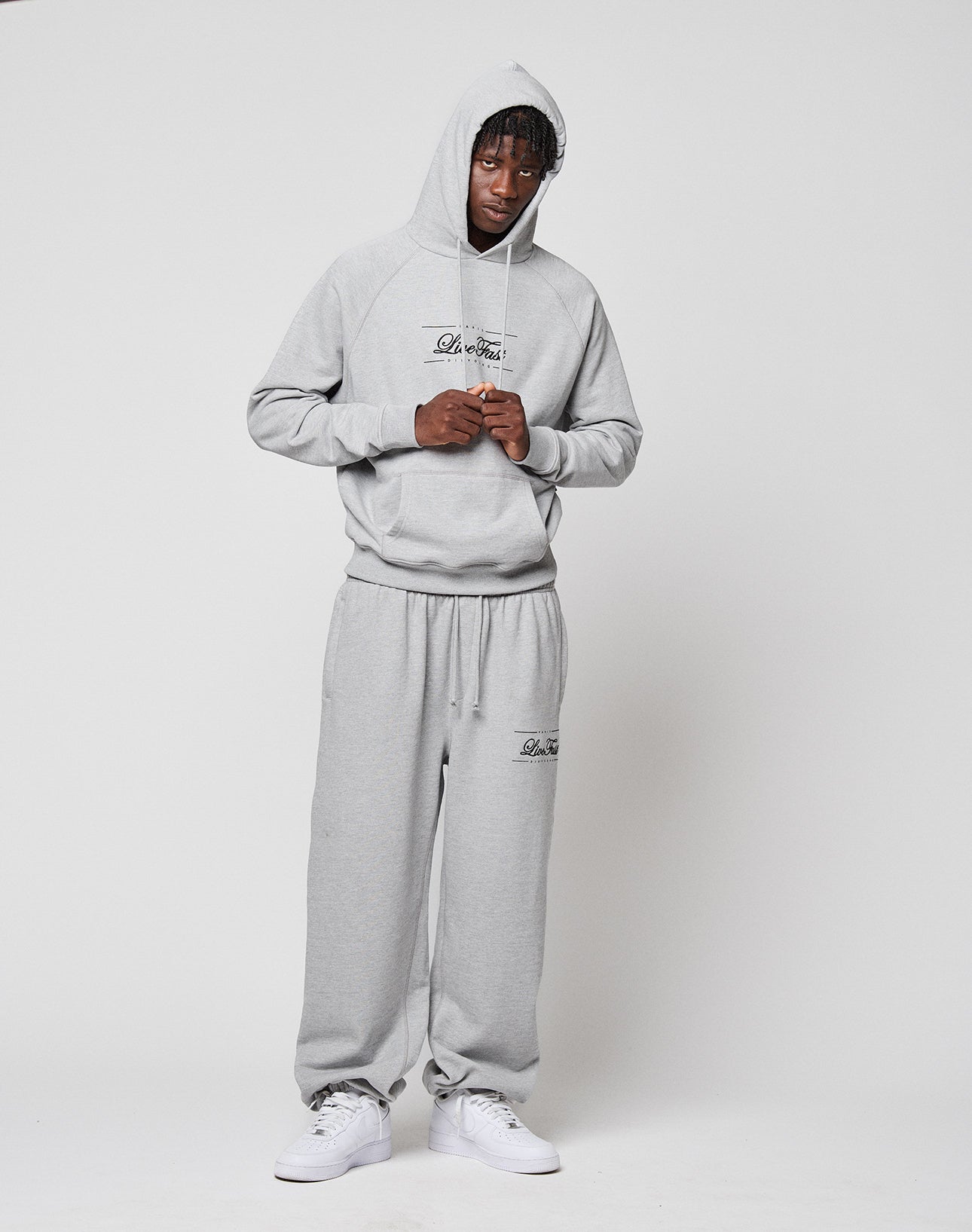 A person in LIVE FAST DIE YOUNG's light gray French Terry Wide Cuff Jogger sweatpants and matching hoodie with elastic waistband stands facing forward against a plain white background, hood up, hands adjusting the collar, wearing white sneakers.