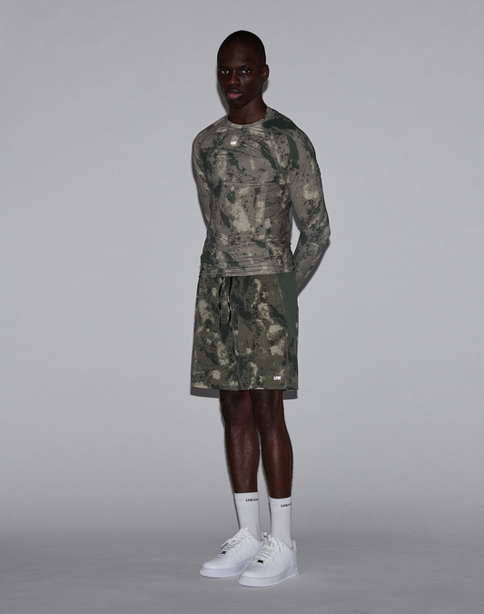 A man stands against a plain gray background, wearing the LIVE FAST DIE YOUNG LFDY Performance Slim Longsleeve, matching camouflage shorts, white socks, and white sneakers. He is facing forward with his arms by his sides.