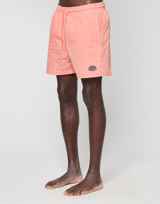 A person stands barefoot against a white background wearing LIVE FAST DIE YOUNG’s light pink Live Fast Swim Shorts, featuring an elastic waistband, side pockets, and a small logo above the left hem.