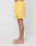 A person stands sideways, barefoot, in regular fit yellow Live Fast Swim Shorts by LIVE FAST DIE YOUNG, featuring a wide elastic waistband, pockets, and a small patch on the left leg, set against a plain white background.