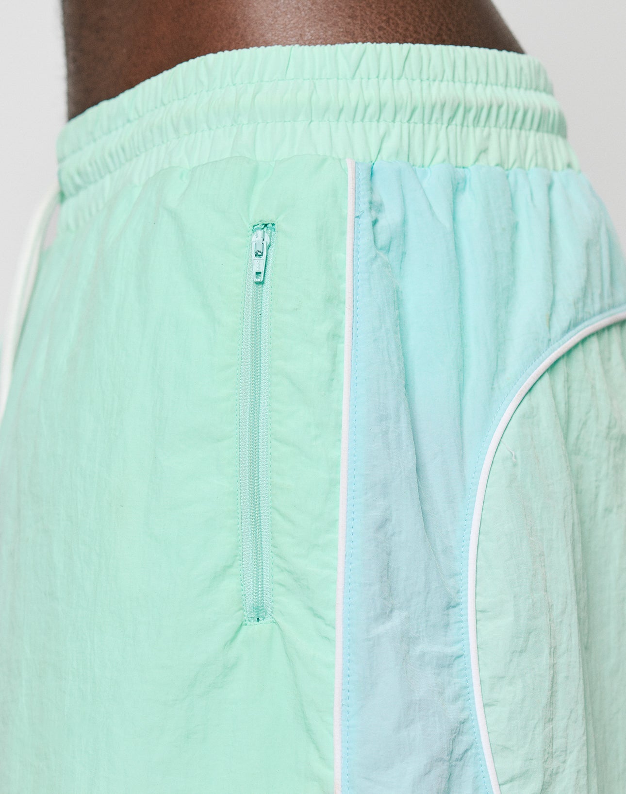 Close-up of a person wearing LIVE FAST DIE YOUNG Beach Shorts in mint green and light blue color-block, featuring zip pockets, elastic waistband, and white piping for a comfortable regular fit.