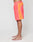 A person stands barefoot in profile against a white background, wearing LIVE FAST DIE YOUNG Beach Shorts in coral and orange color block with zippered side pockets, wide elastic waistband, white piping, and a drawstring waist.