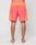 A person stands barefoot, facing away, wearing LIVE FAST DIE YOUNG Beach Shorts in bright orange and coral with white piping and a wide elastic waistband, set against a plain light background.