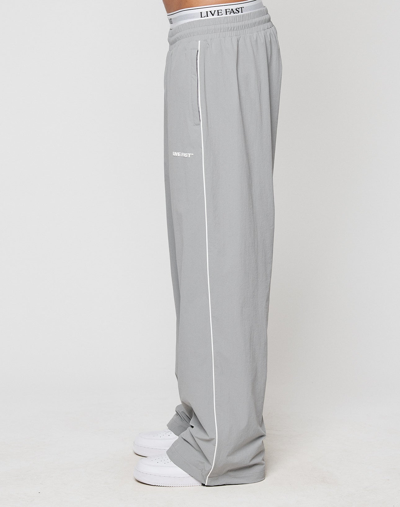 A person stands against a white background wearing LIVE FAST DIE YOUNG LFDY Ripstop Trackpants in loose, wide fit light gray with white piping and LIVE FAST Silikonprint near the pocket, paired with white sneakers.