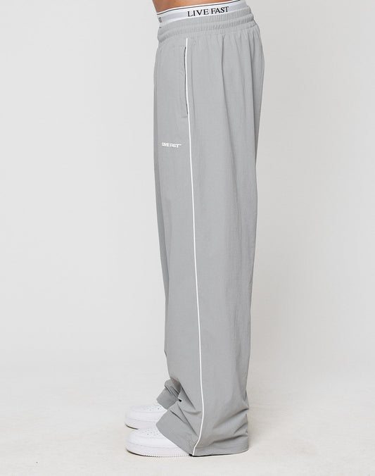 A person stands against a white background wearing LIVE FAST DIE YOUNG LFDY Ripstop Trackpants in loose, wide fit light gray with white piping and LIVE FAST Silikonprint near the pocket, paired with white sneakers.
