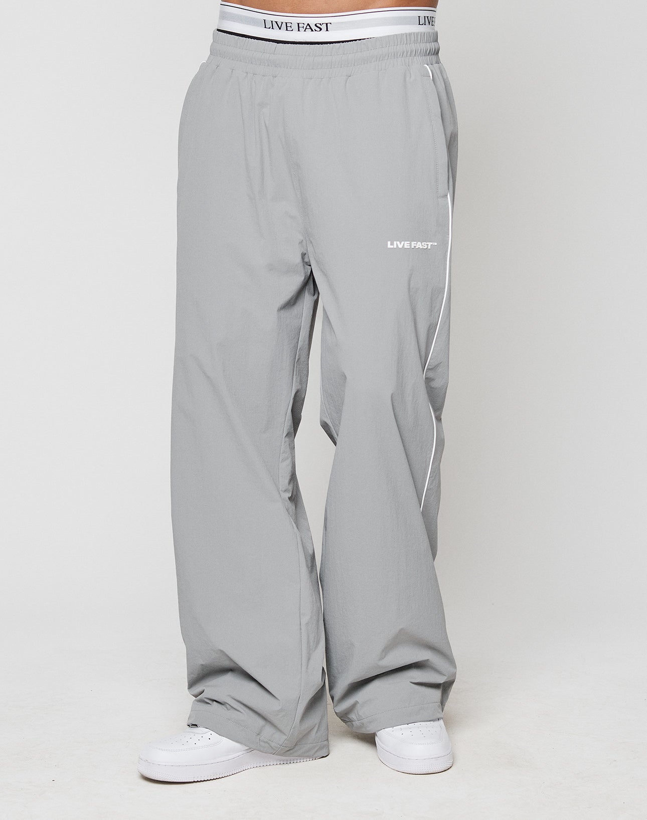 A person wears LIVE FAST DIE YOUNG LFDY Ripstop Trackpants in loose-fitting light gray with wide fit, silicone print on waistband and upper thigh, and elastic waistband. Paired with white sneakers against a plain white background.