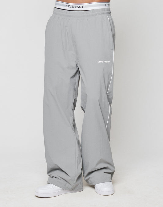 A person wears LIVE FAST DIE YOUNG LFDY Ripstop Trackpants in loose-fitting light gray with wide fit, silicone print on waistband and upper thigh, and elastic waistband. Paired with white sneakers against a plain white background.