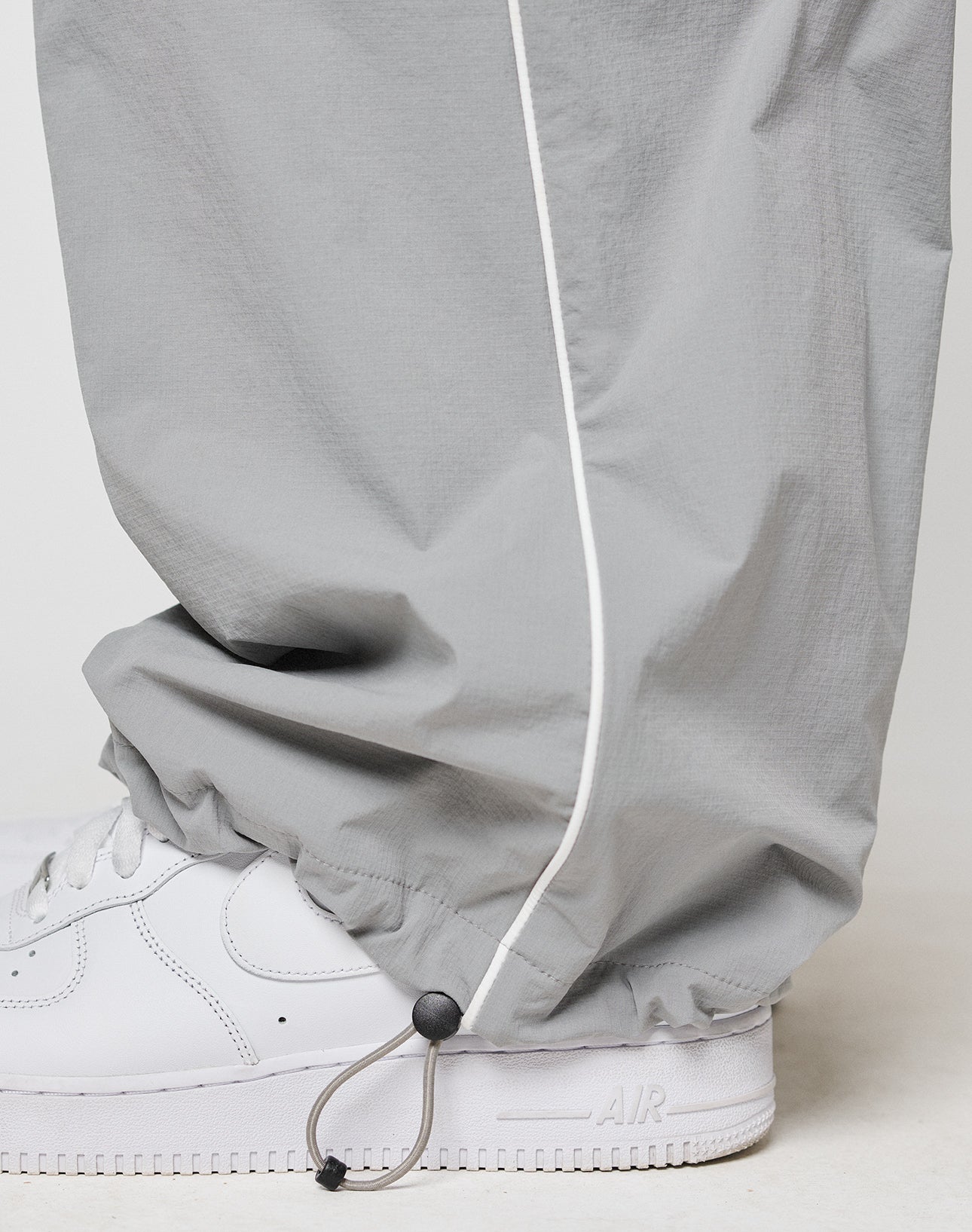 A close-up of a person wearing LIVE FAST DIE YOUNG LFDY Ripstop Trackpants in light gray with an elastic waistband, white piping, ankle drawstring, paired with white Nike Air Force 1 sneakers.