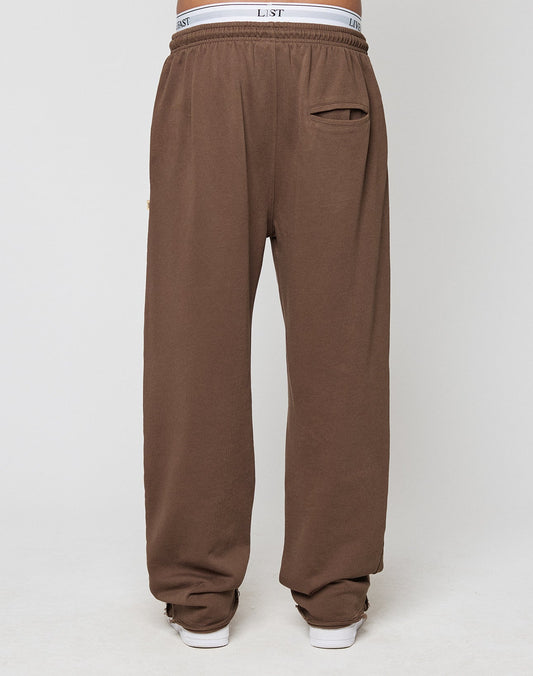 A person wears LIVE FAST DIE YOUNG’s LFDY Lightweight Jogger—loose-fitting brown French Terry sweatpants with an elastic waistband, visible white underwear, and a small back pocket—standing on a light background.