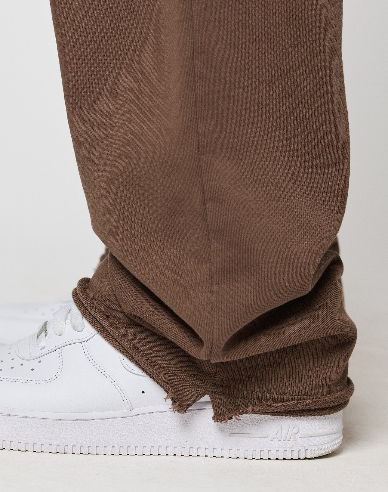 A close-up of lower legs and feet wearing LIVE FAST DIE YOUNG’s LFDY Lightweight Jogger in brown, featuring an oversize fit and unfinished hems, paired with clean white Nike Air Force 1 sneakers.
