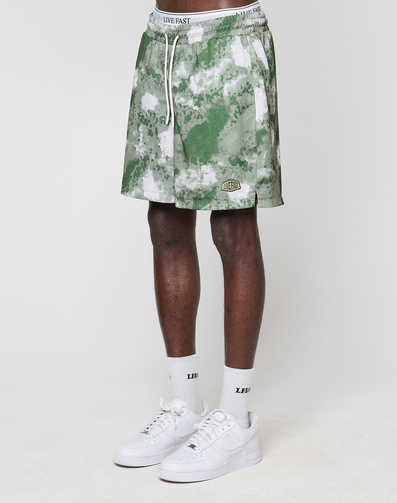 A person wears green and white LFDY Mesh Shorts by LIVE FAST DIE YOUNG (Regular Fit, elastic waistband), white LIVE FAST socks, and white Nike sneakers. Only their legs and lower torso are shown against a plain white background. | model