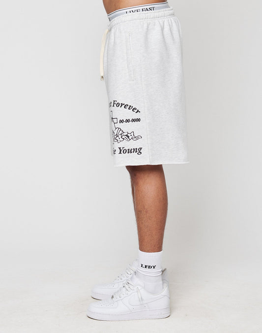 A person stands sideways in LFDY Praying Shorts by LIVE FAST DIE YOUNG, light gray with black graphic text and illustration on the left leg, featuring seitliche Eingrifftaschen, Elastikbund, and paired with white crew socks and sneakers. Plain white background.