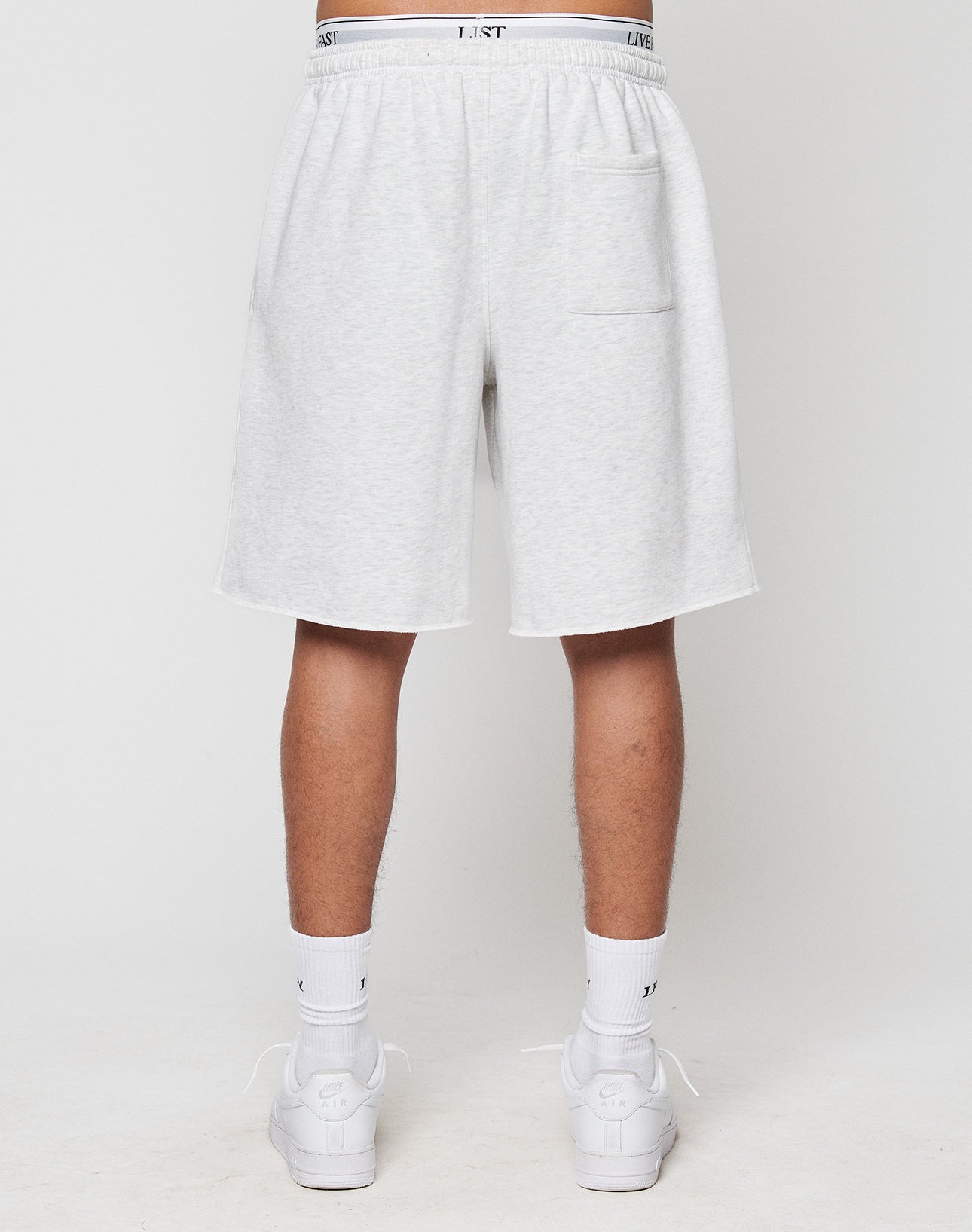 A person stands with their back to the camera, showing only legs and lower torso in LFDY Praying Shorts by LIVE FAST DIE YOUNG—light gray with elastic waistband and side pockets—paired with white crew socks and sneakers against a plain background.