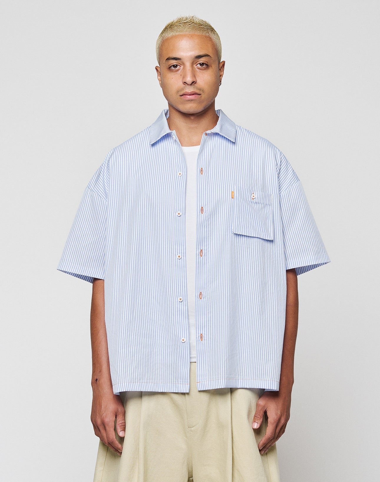 A person with short blonde hair wears the LFDY Striped Boxy Shirt by LIVE FAST DIE YOUNG in light blue and white stripes with a chest pocket and subtle logo, paired with beige wide-leg pants, against a plain light gray background. | model