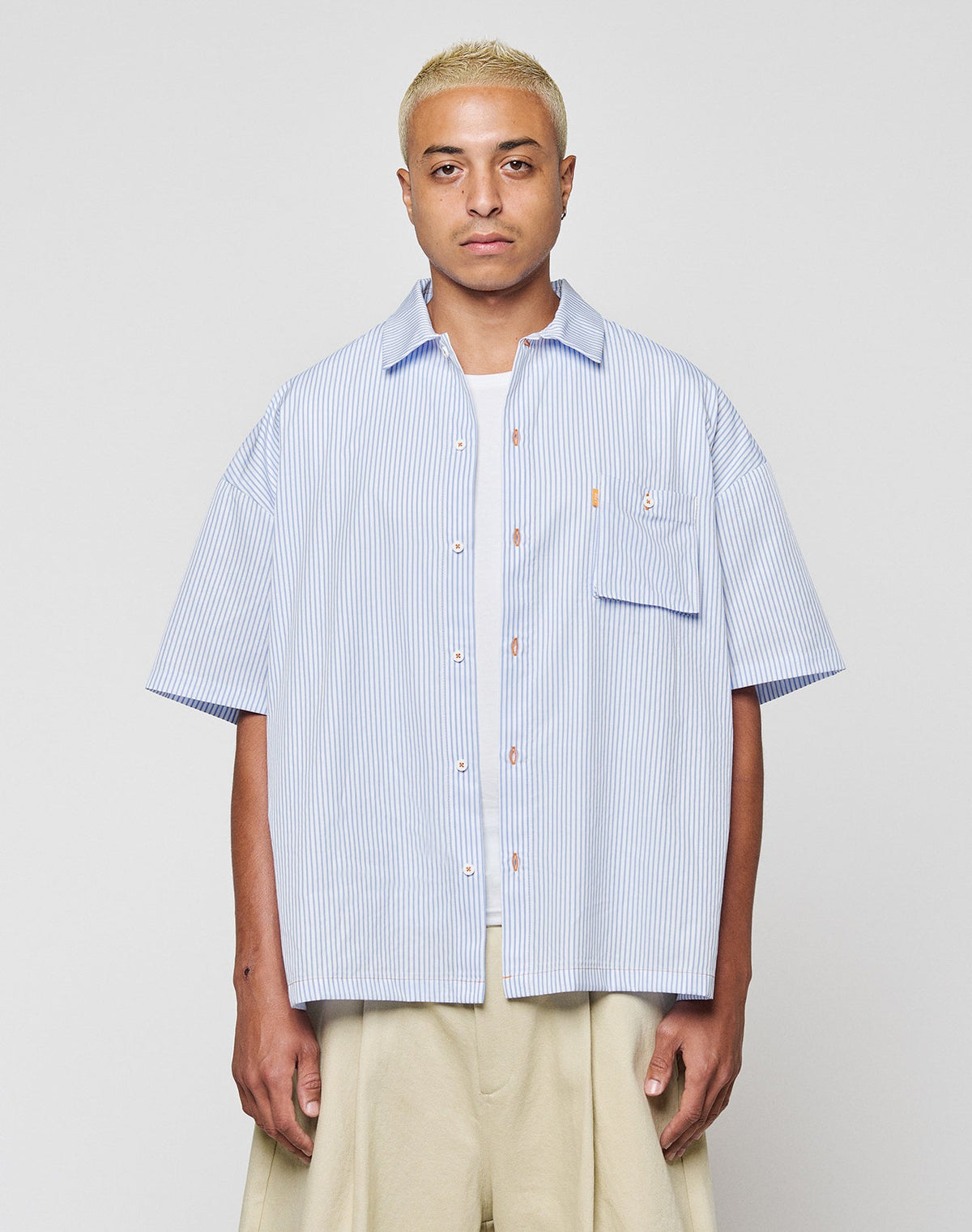 A person with short blonde hair wears the LFDY Striped Boxy Shirt by LIVE FAST DIE YOUNG in light blue and white stripes with a chest pocket and subtle logo, paired with beige wide-leg pants, against a plain light gray background. | model