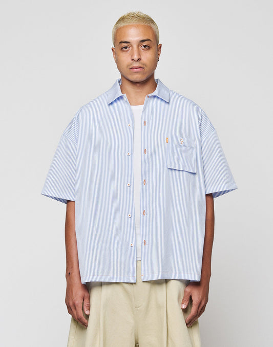 A person with short blonde hair wears the LFDY Striped Boxy Shirt by LIVE FAST DIE YOUNG in light blue and white stripes with a chest pocket and subtle logo, paired with beige wide-leg pants, against a plain light gray background. | model