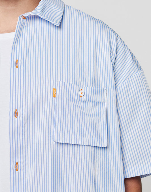 A person wears the LIVE FAST DIE YOUNG LFDY Striped Boxy Shirt with light blue and white stripes, featuring a Blasebalg Tasche with orange buttons and subtle logo stitch, layered over a white undershirt. Only the upper torso is visible.