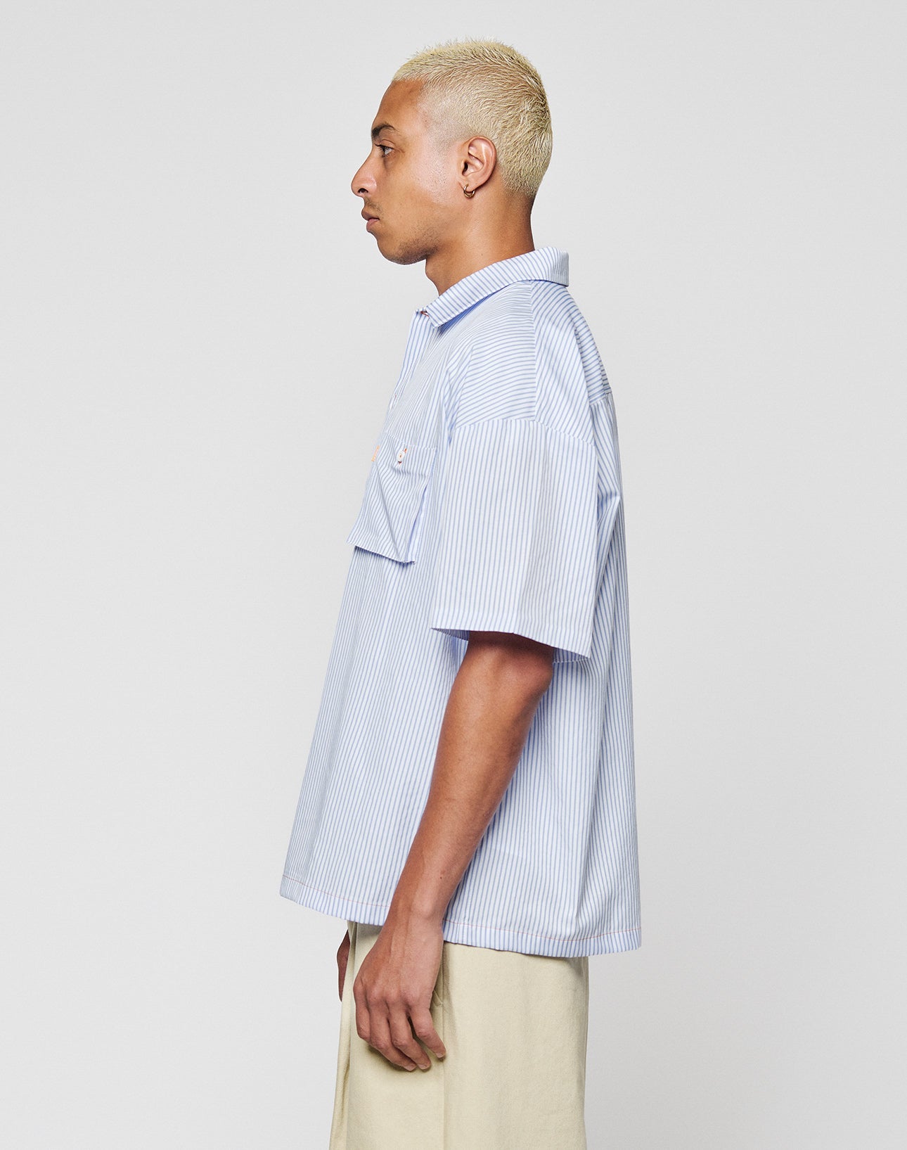 A person with short, bleached blond hair is shown in profile, wearing the LIVE FAST DIE YOUNG LFDY Striped Boxy Shirt in light blue and white stripes and light beige pants, standing against a plain light gray background.