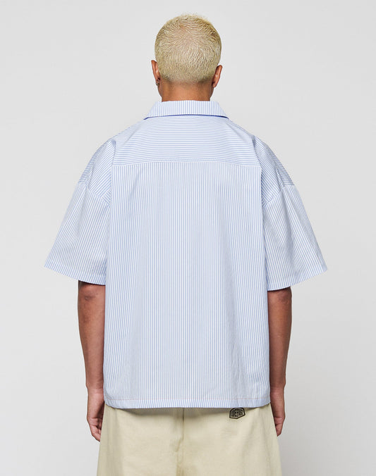 A person with short bleached blond hair is shown from behind, wearing the LIVE FAST DIE YOUNG LFDY Striped Boxy Shirt with blue and white vertical stripes and cream pants, featuring a subtle logo-stitch detail against a plain background.