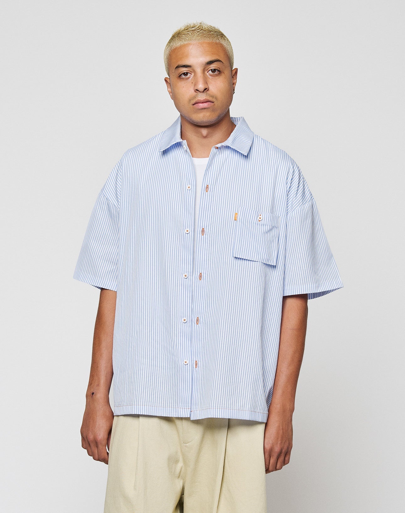 A person with short blond hair wears the LIVE FAST DIE YOUNG LFDY Striped Boxy Shirt in light blue and white, featuring a subtle logo stitch, paired with beige pants. They stand against a plain light gray background.