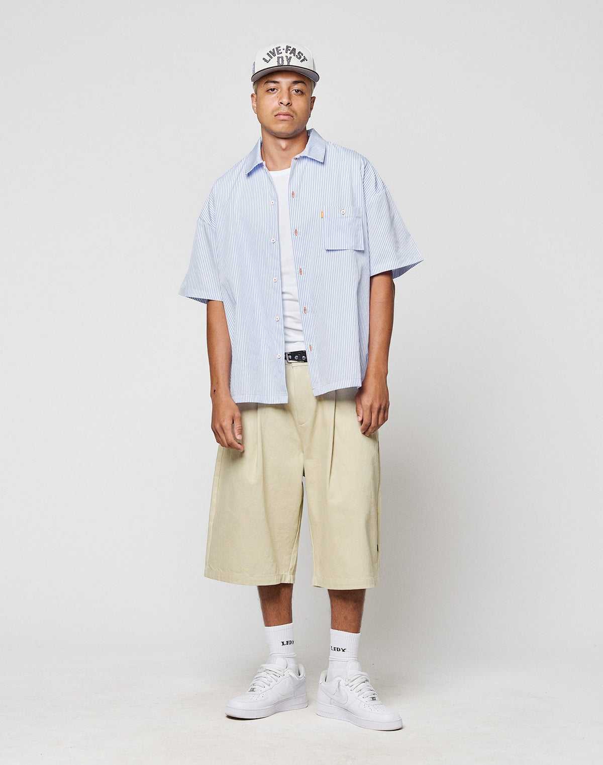 A man stands against a plain background wearing a LIVE FAST DIE YOUNG LFDY Striped Boxy Shirt over a white T-shirt, beige wide-leg shorts, white socks, white sneakers, and a gray cap with Logo-Stick text. A Blasebalg Tasche completes the look.