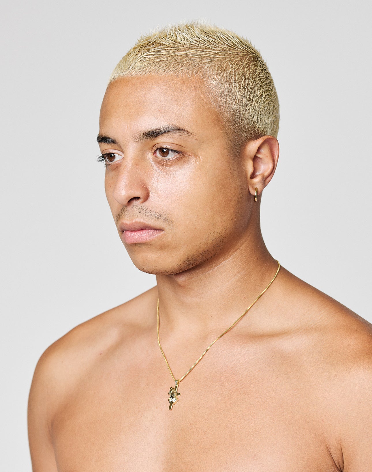 A person with short, bleached blonde hair and medium skin tone looks to the side. They are shirtless, wearing a LIVE FAST DIE YOUNG LFDY Praying Chain necklace with pendant and a small gold hoop earring. Background is plain and light-colored.