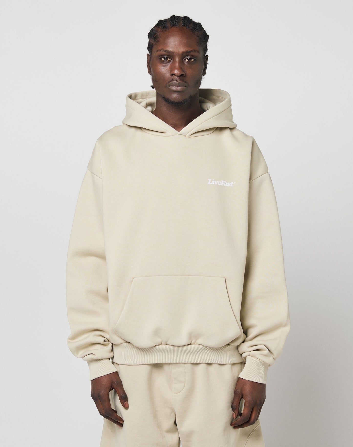 A person with braided hair wears the LIVE FAST DIE YOUNG Basic 360 Hooded in beige, styled with matching oversize sweatpants. A small white LiveFast logo appears on the chest. The background is plain white.