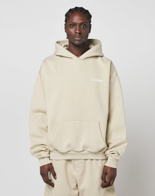 A person with braided hair wears the LIVE FAST DIE YOUNG Basic 360 Hooded in beige, styled with matching oversize sweatpants. A small white LiveFast logo appears on the chest. The background is plain white.