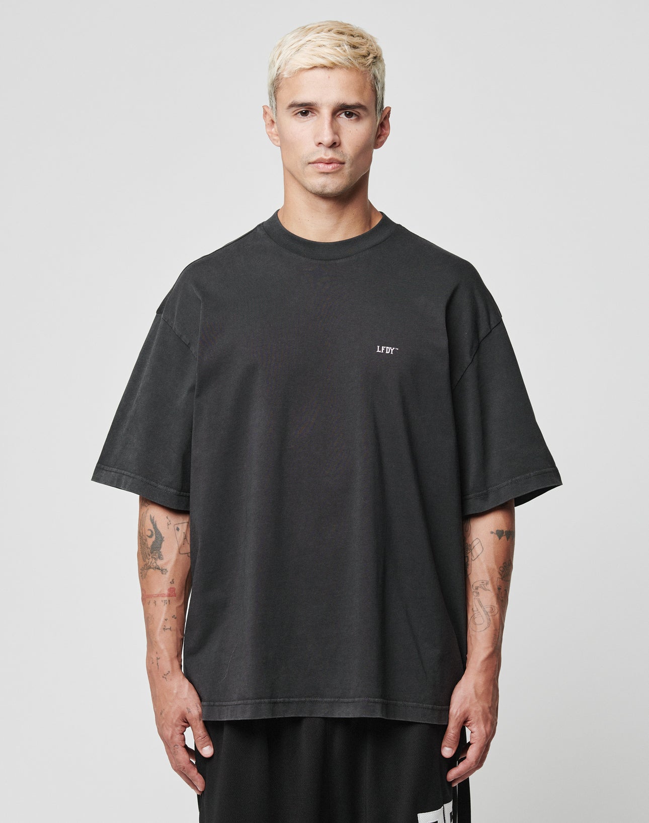 A man with short bleached blonde hair wears the black LFDY Flip Phone Tee by LIVE FAST DIE YOUNG and black shorts, standing against a plain gray background, looking directly at the camera with a neutral expression.