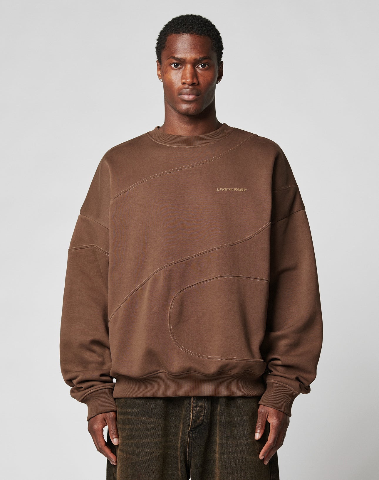 A man with short curly hair and a neutral expression wears the oversize LFDY Wavy Sweater by LIVE FAST DIE YOUNG—a brown Baumwolle-Polyester blend sweatshirt with stitched curves and LIVE IN FAST on the chest. Plain light gray background.