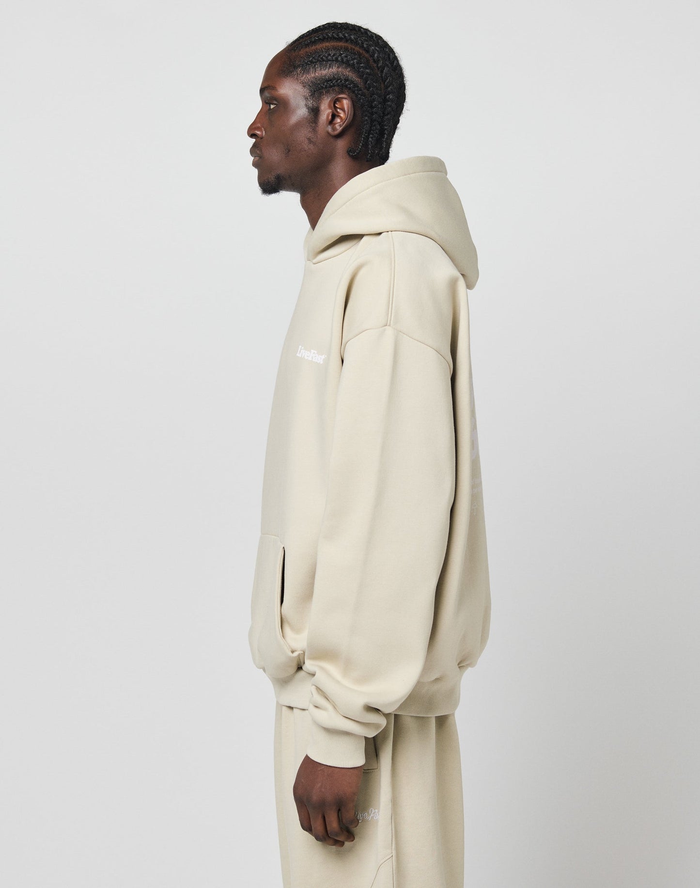 A person in profile stands against a white background, wearing the LIVE FAST DIE YOUNG Basic 360 Hooded—an oversize beige hoodie and matching sweatpants crafted from soft Baumwolle and durable polyester, with hands relaxed by their sides.