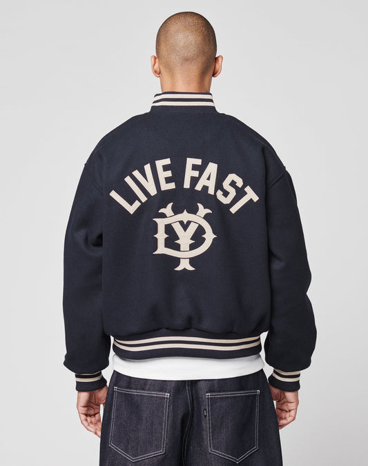 A person with a shaved head wears the LIVE FAST DIE YOUNG Live Fast College Jacket featuring beige trim, a DY-Reißverschluss Puller, large beige LIVE FAST text, a stylized LY logo on the back, and a classic regular fit. | model