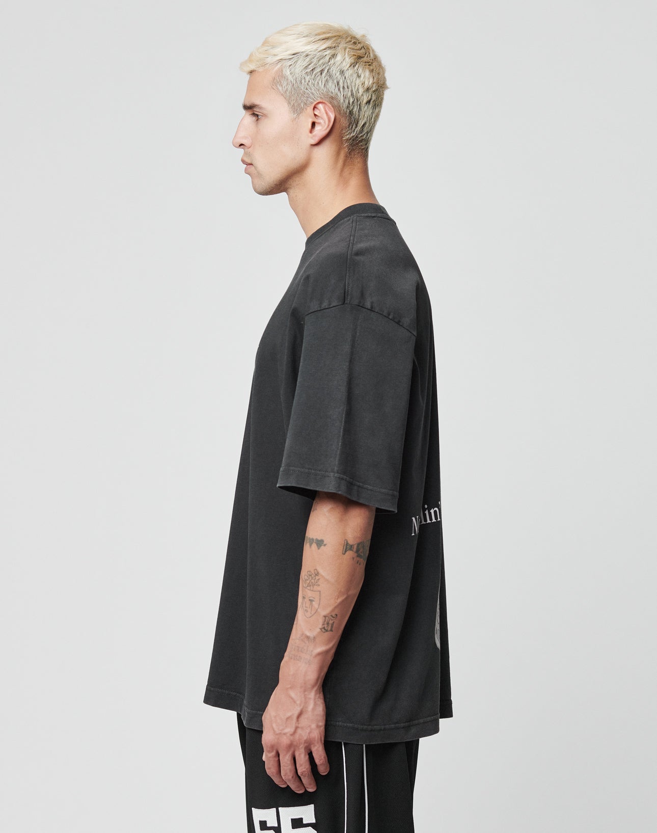 A man with short, bleached blond hair and tattooed arms stands in profile wearing a LIVE FAST DIE YOUNG LFDY Flip Phone Tee and black pants with white stripes and numbers against a plain light gray background.