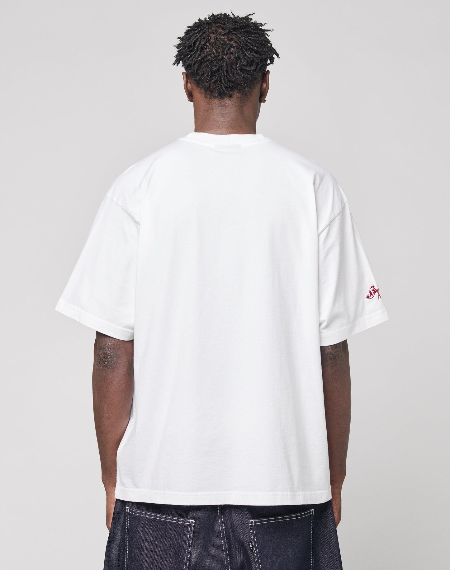 A person with short, textured hair stands with their back to the camera, wearing the oversize fit Live Fast Stallion Tee by LIVE FAST DIE YOUNG and dark jeans, against a light gray background.