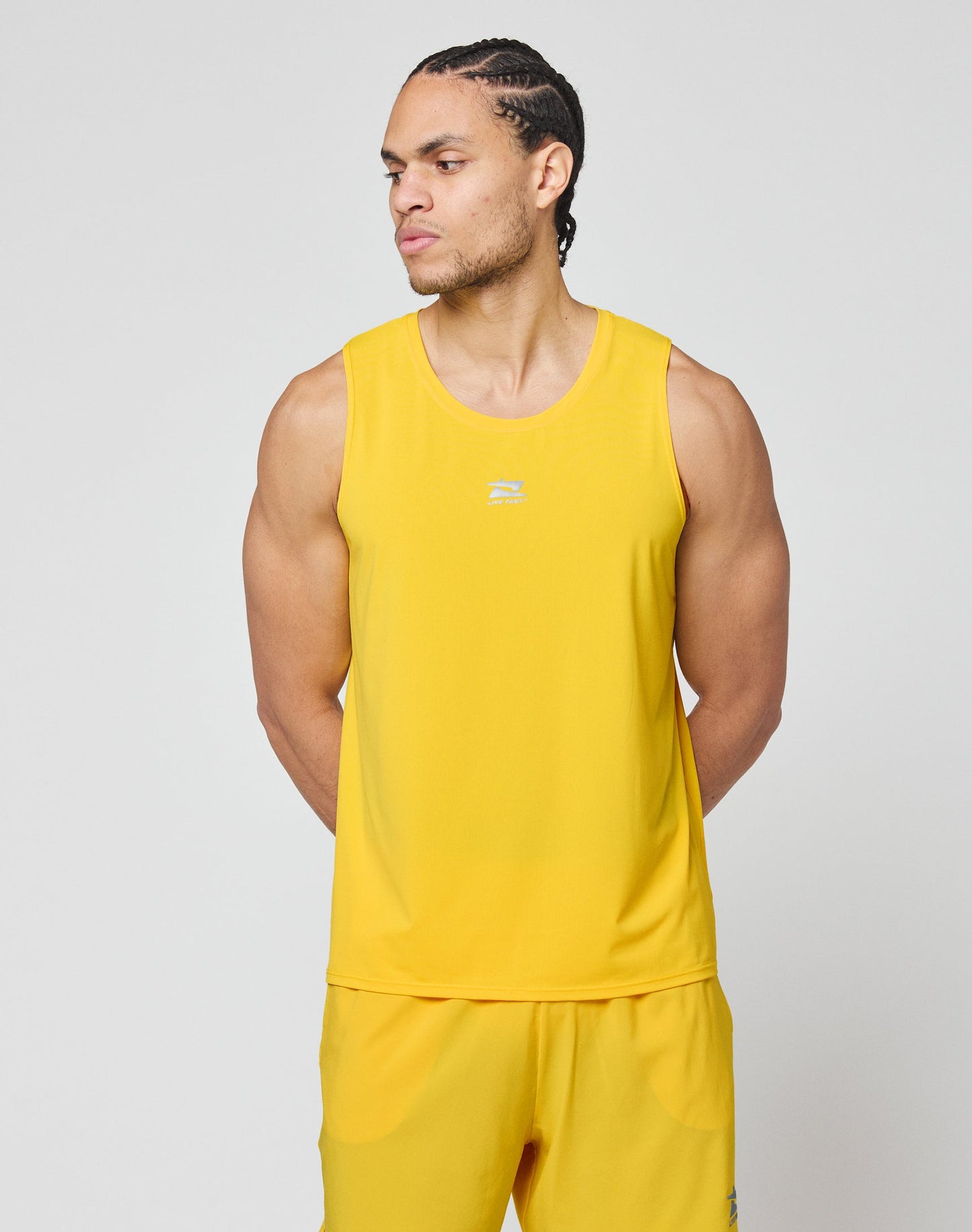 A man with braided hair stands in profile, wearing a slim fit, sleeveless yellow Live Fast Performance Tank and matching yellow shorts by LIVE FAST DIE YOUNG against a plain light gray background.