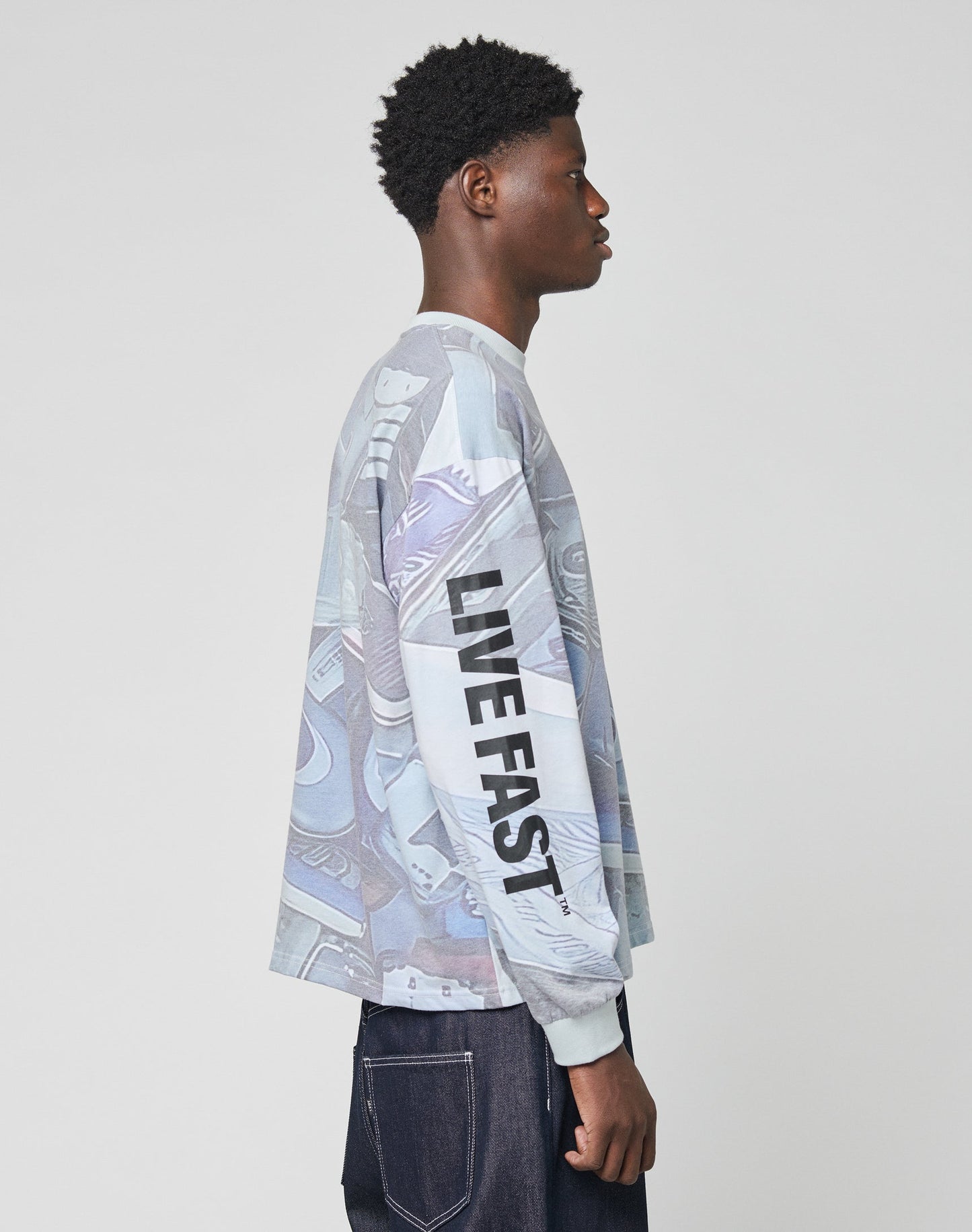A man stands in profile wearing the LFDY Stolen Phones Longsleeve by LIVE FAST DIE YOUNG, featuring auffälligem Ärmelprint with bold LIVE FAST lettering on the sleeve, paired with dark vintage-wash jeans against a light background.