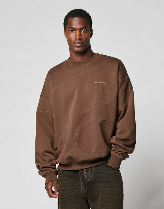 A man stands against a light gray background, wearing the LFDY Wavy Sweater by LIVE FAST DIE YOUNG—an oversized brown sweatshirt with subtle stitching, “LIVE IN PART” on the chest, and paired with dark jeans.