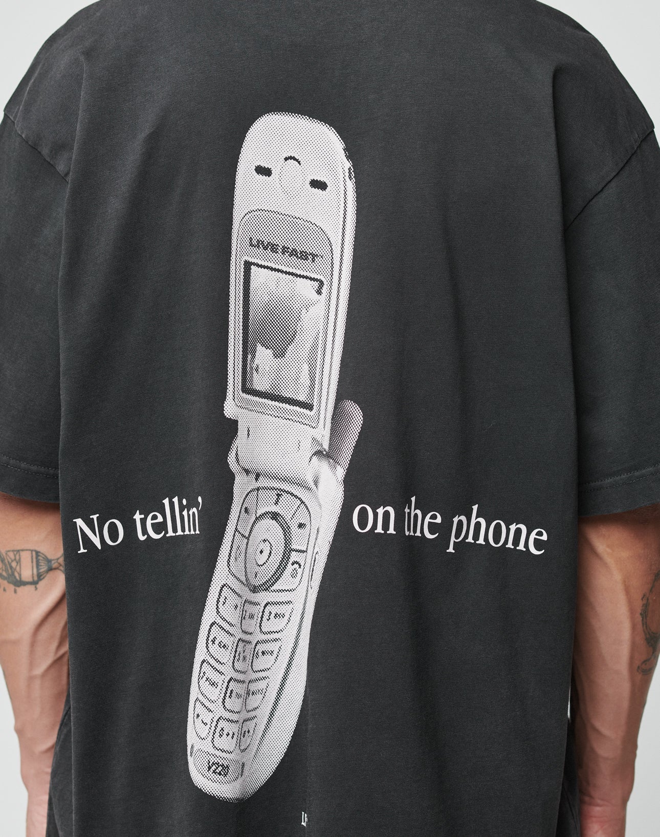 Someone wears the LFDY Flip Phone Tee by LIVE FAST DIE YOUNG, an oversize black Baumwolle t-shirt with a large old flip phone graphic and “No tellin on the phone” printed on the back.