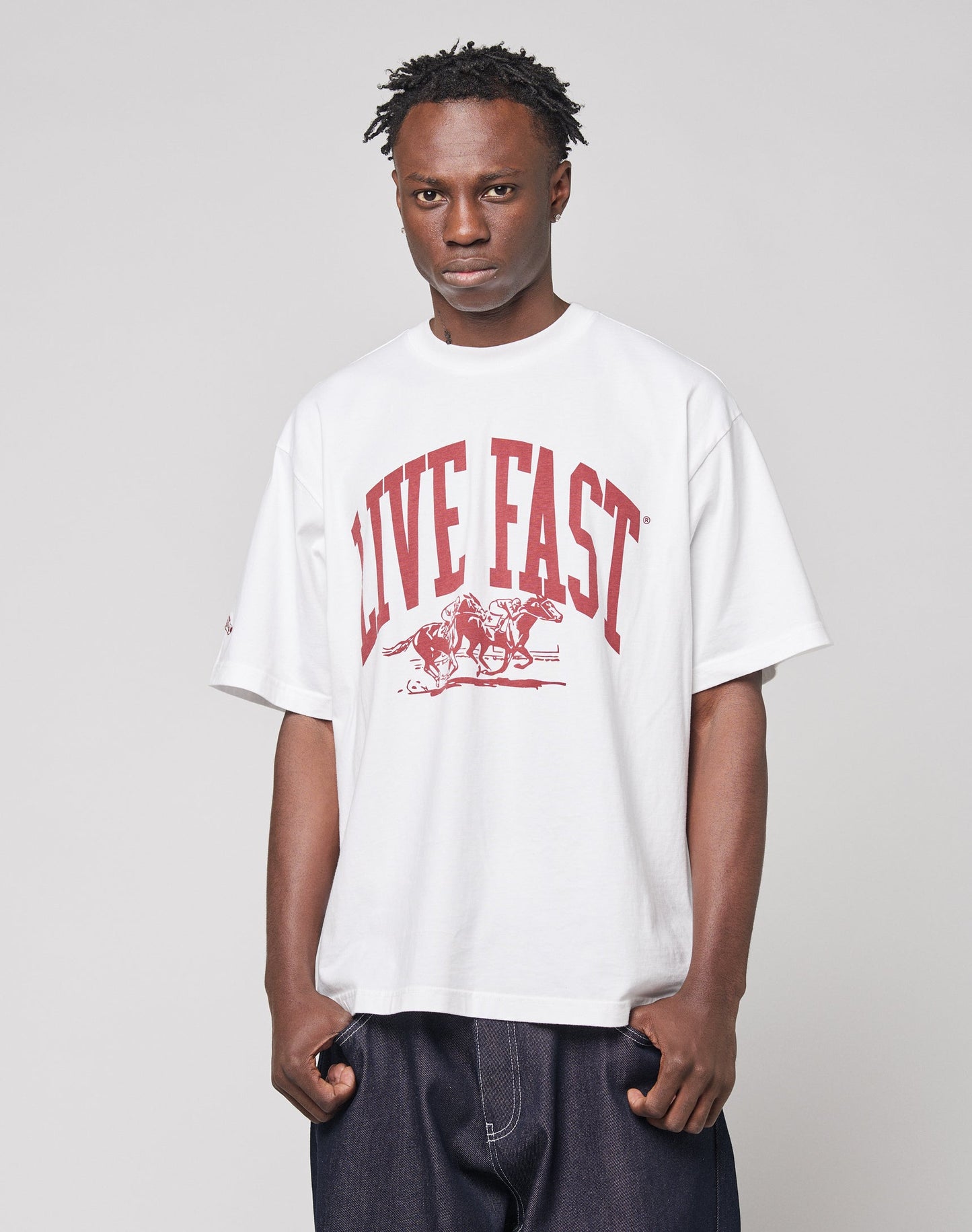 A man wears the LIVE FAST DIE YOUNG Live Fast Stallion Tee—an oversized white cotton T-shirt with a bold red “LIVE FAST” print and a speeding horse and rider graphic—paired with dark, loose jeans against a plain background.