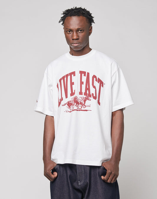 A man wears the LIVE FAST DIE YOUNG Live Fast Stallion Tee—an oversized white cotton T-shirt with a bold red “LIVE FAST” print and a speeding horse and rider graphic—paired with dark, loose jeans against a plain background.