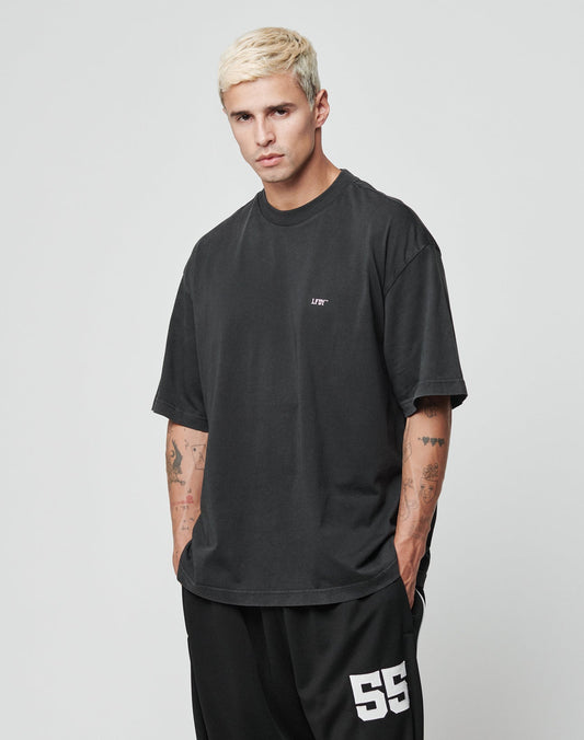 A young man with short bleached blond hair wears the LIVE FAST DIE YOUNG LFDY Flip Phone Tee in an oversized black cotton fit with small white text and black shorts numbered 55, standing with hands in pockets against a plain light background.