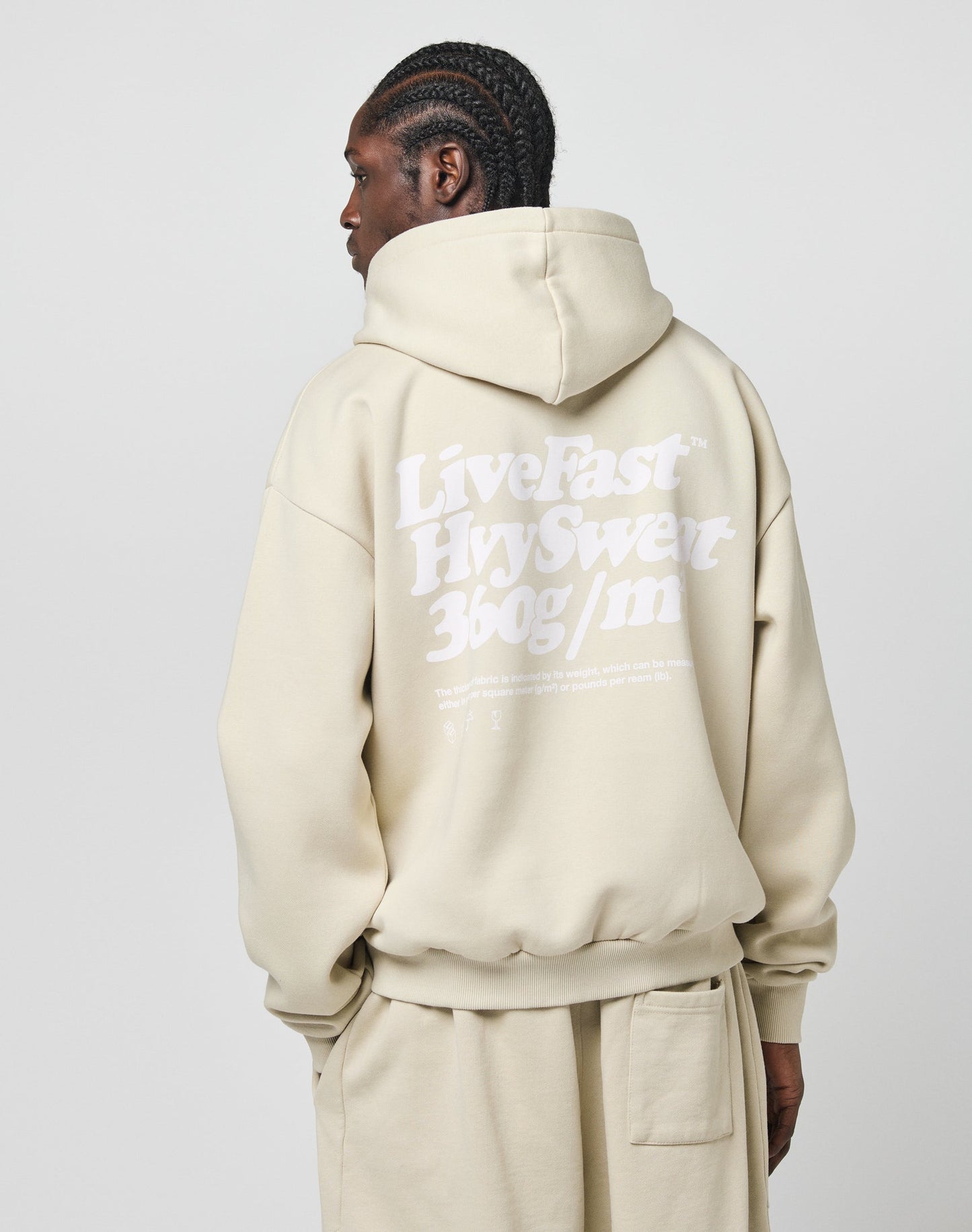 Wearing the Basic 360 Hooded by LIVE FAST DIE YOUNG, a person with braided hair, facing away from the camera against a light background, sports an oversize beige hoodie with Live Fast in white text on the back.