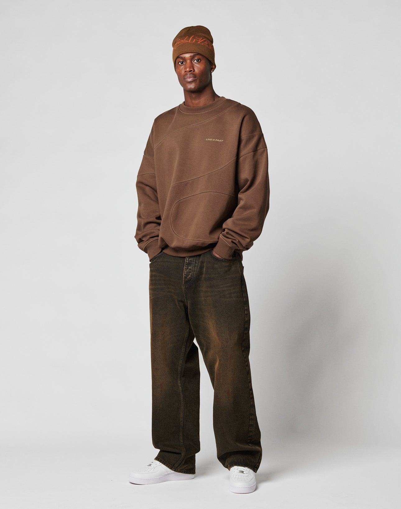 A man stands against a plain background wearing the LIVE FAST DIE YOUNG LFDY Wavy Sweater in brown, paired with a brown knit beanie, loose dark brown jeans, and white sneakers. His hands are in his sweater pockets as he faces the camera.